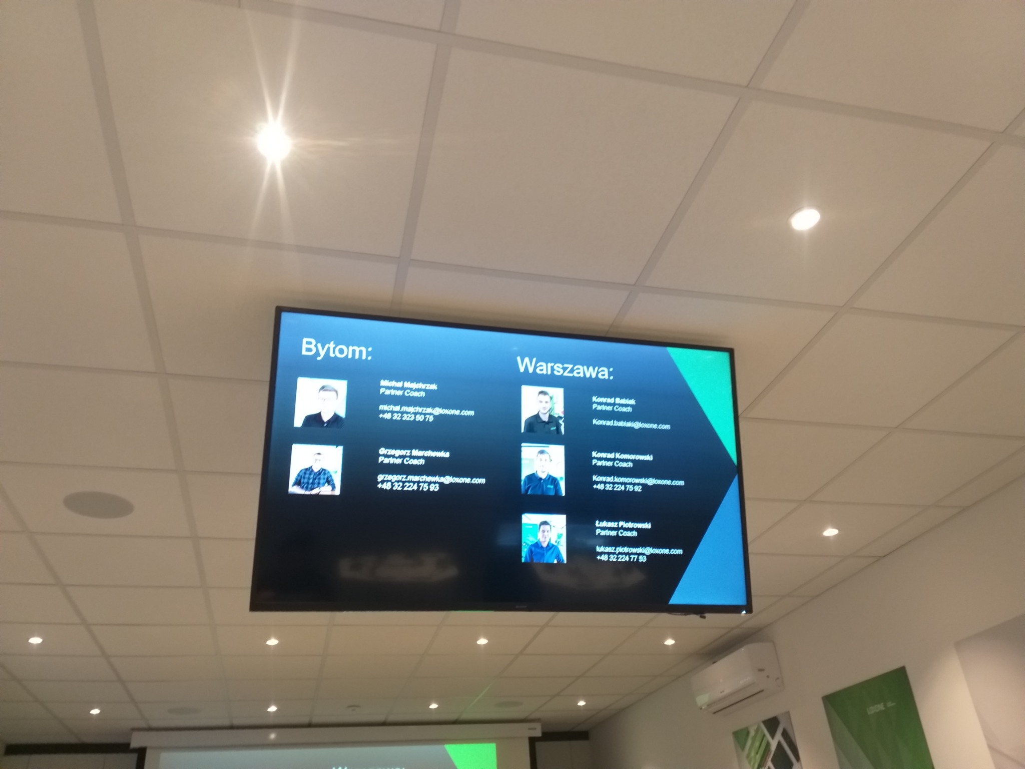Ekran wyświetlający listę partnerów Loxone z Bytomia i Warszawy, z ich zdjęciami i danymi kontaktowymi, zawieszony pod sufitem podwieszanym z oświetleniem punktowym.