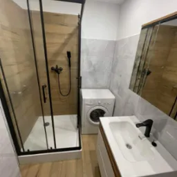 Nowoczesna łazienka po remoncie: kabina prysznicowa z czarnymi profilami, umywalka z szafką i czarną armaturą, pralka oraz płytki imitujące drewno i marmur.