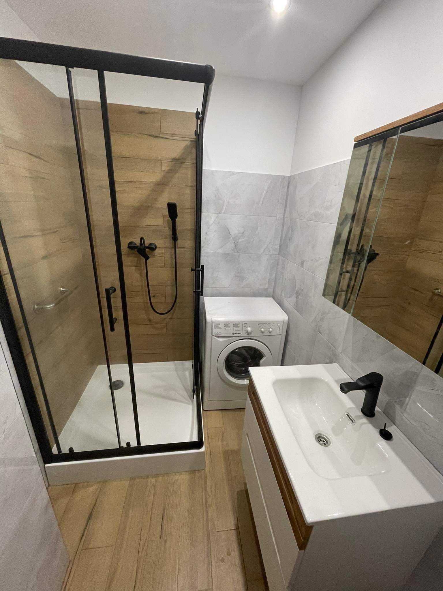 Nowoczesna łazienka po remoncie: kabina prysznicowa z czarnymi profilami, umywalka z szafką i czarną armaturą, pralka oraz płytki imitujące drewno i marmur.