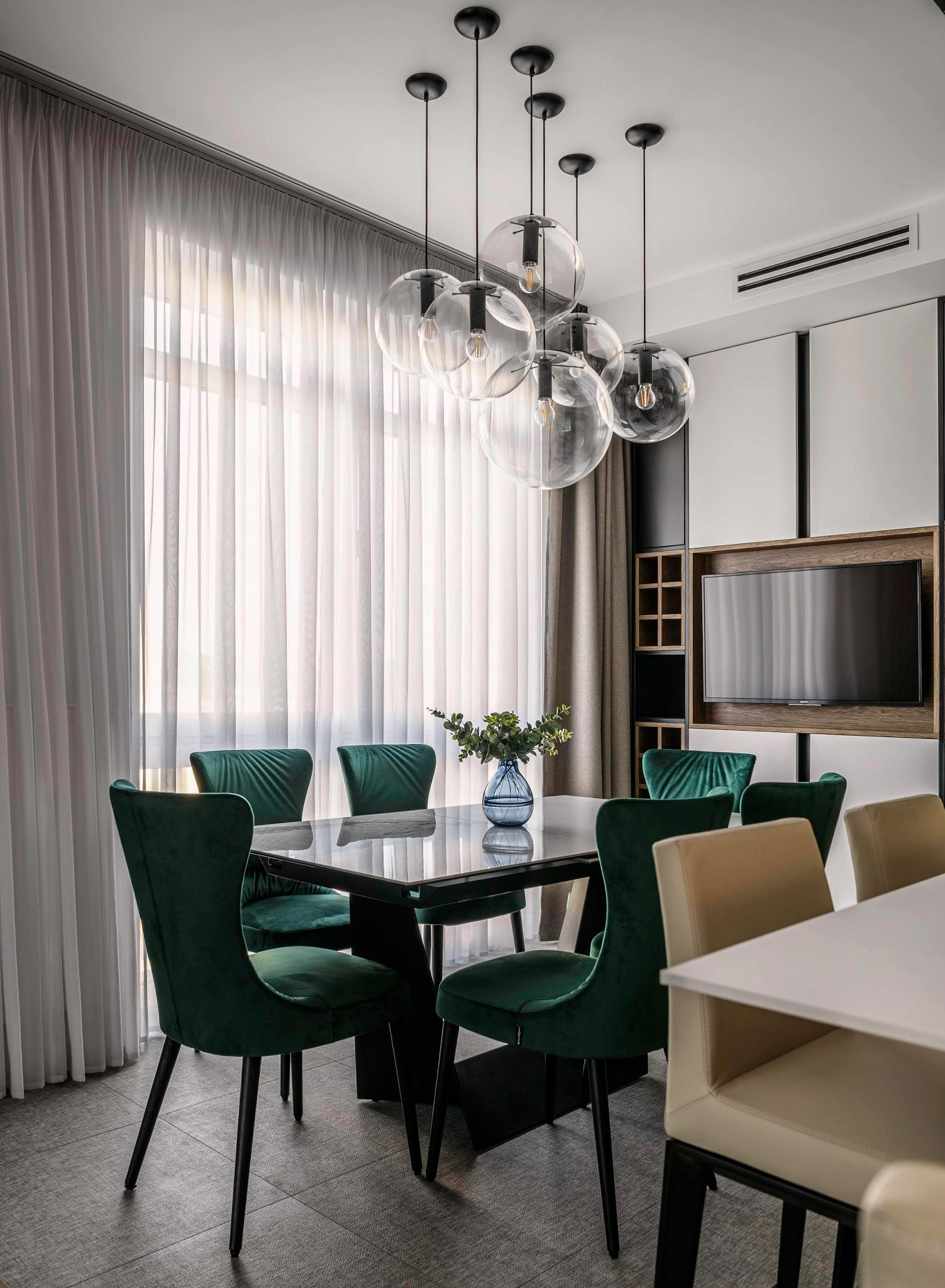 Jadalnia z zasłonami woalowymi, szklanym stołem, zielonymi krzesłami i lampami wiszącymi w kształcie kul.