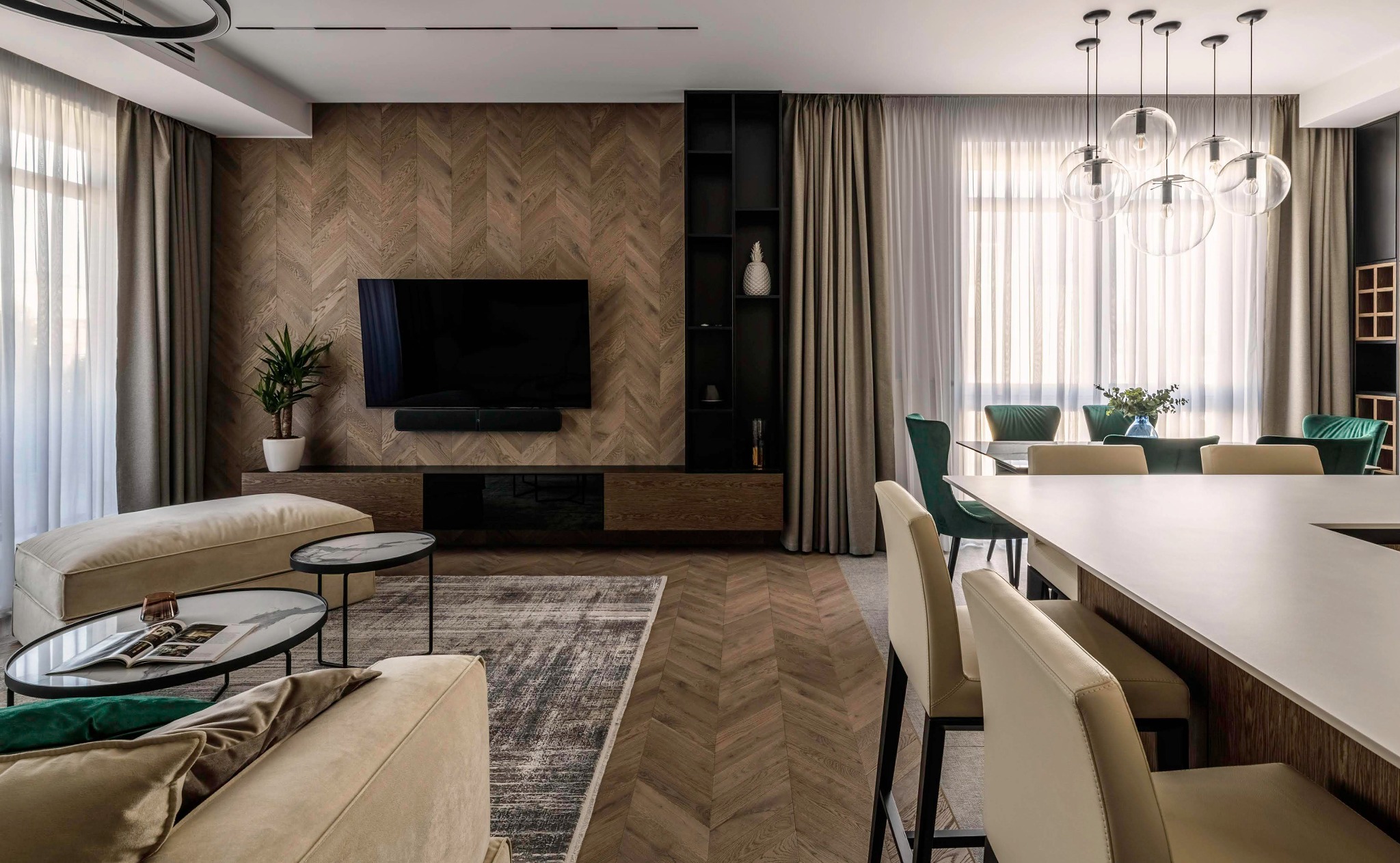 Nowoczesne wnętrze salonu z zasłonami w odcieniach beżu, drewnianą ścianą z telewizorem, szklanymi lampami wiszącymi i elementami wystroju w kolorze zielonym.