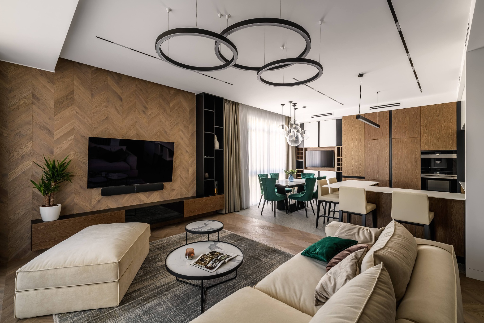 Nowoczesny salon z drewnianą ścianą w jodełkę, okrągłymi lampami sufitowymi, beżową sofą z zielonymi poduszkami, stolikami kawowymi z marmurowymi blatami i zielonymi krzesłami w jadalni w tle.