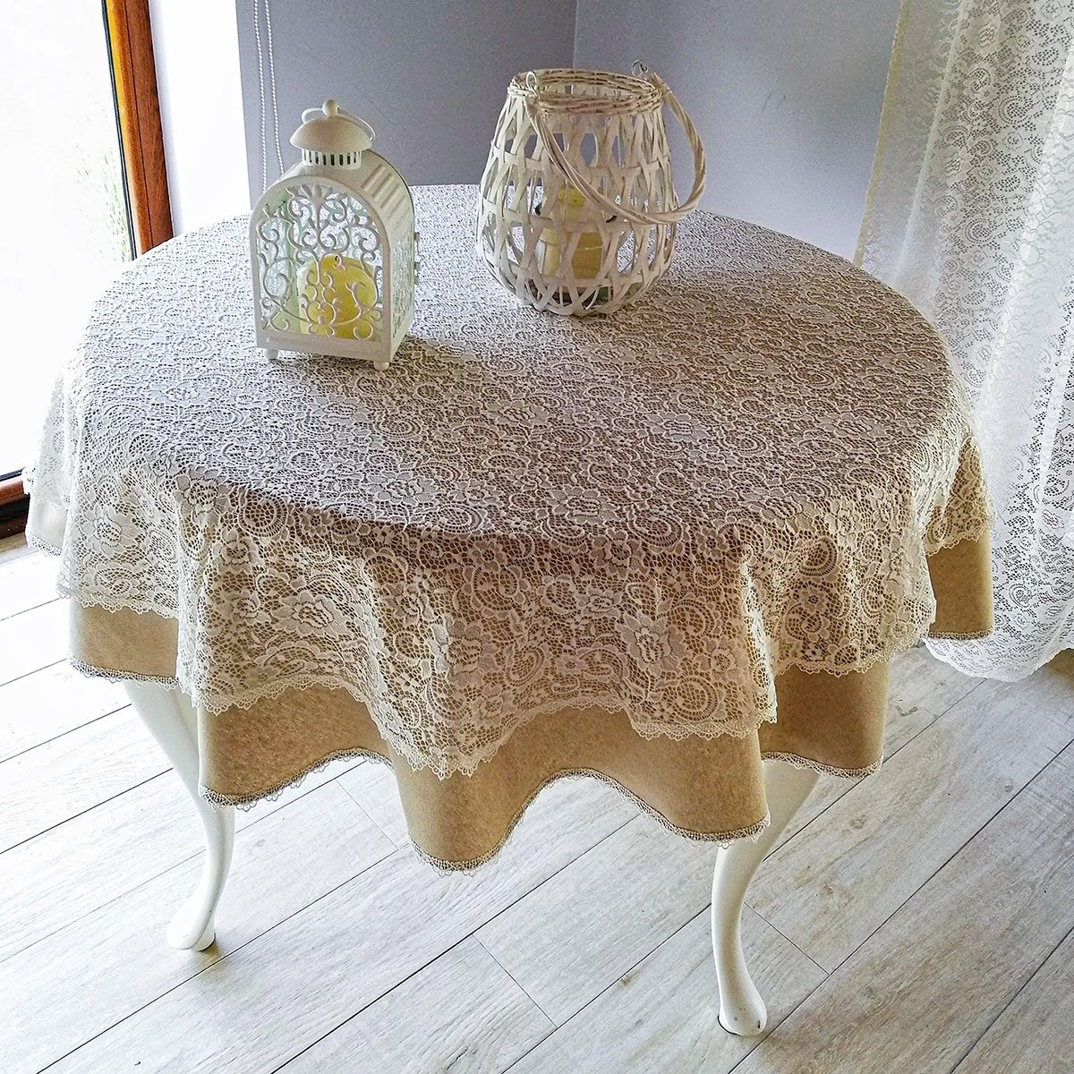 Okrągły stół przykryty beżowym obrusem z koronkową nakładką, dekorowany dwoma lampionami w stylu rustykalnym, na podłodze jasne panele.