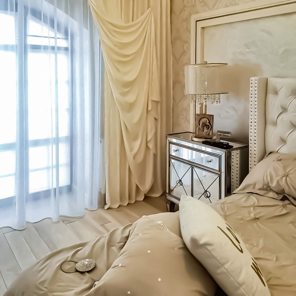 Elegancka sypialnia z beżowymi zasłonami drapowanymi przy oknie, lustrzana szafka nocna z lampką i obrazkiem, łóżko z beżową pościelą i ozdobnymi poduszkami.