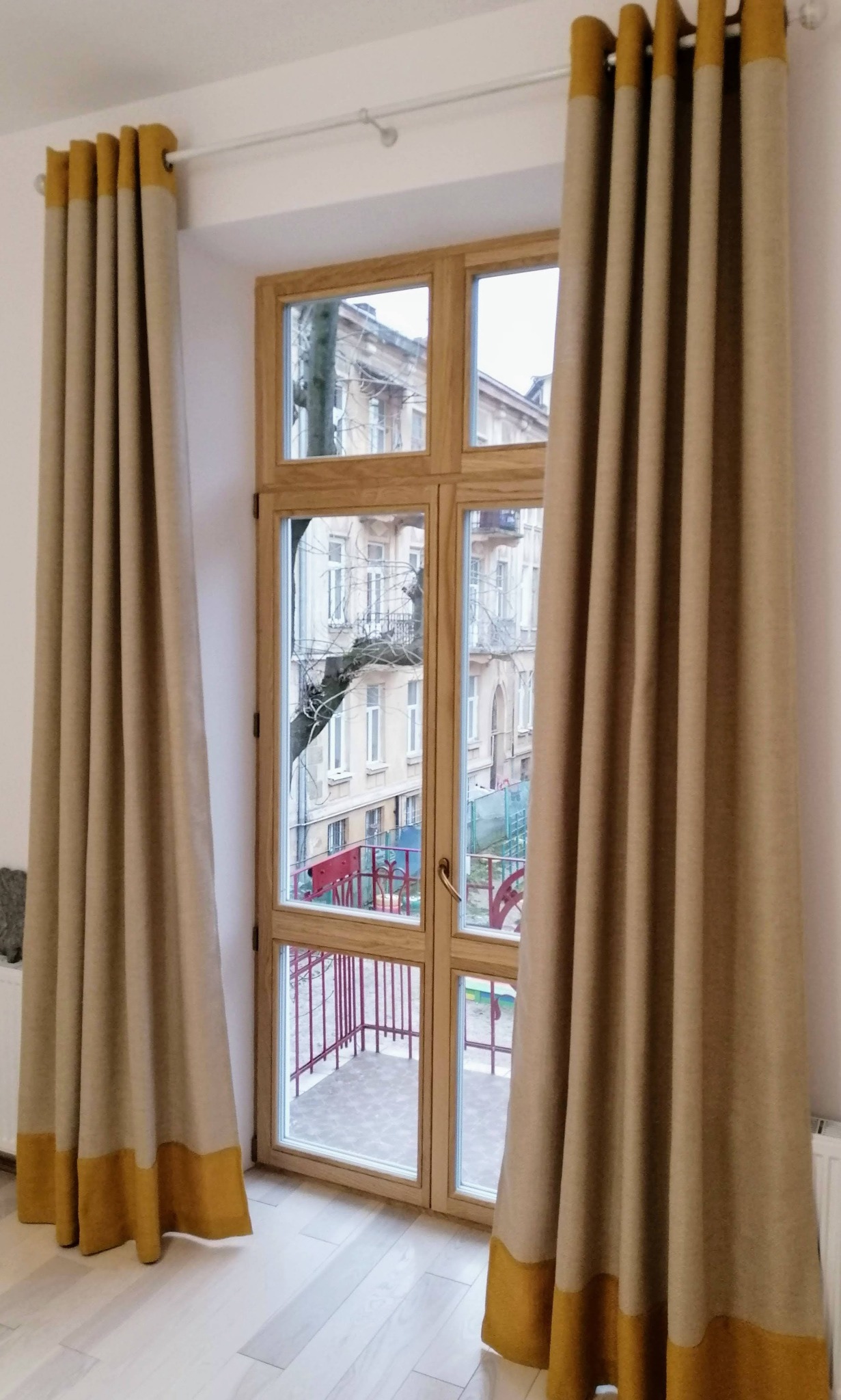 Długie, beżowe zasłony z żółtymi akcentami u góry i na dole, wiszące po obu stronach okna balkonowego z drewnianą ramą, widok na balkon i budynek naprzeciwko.