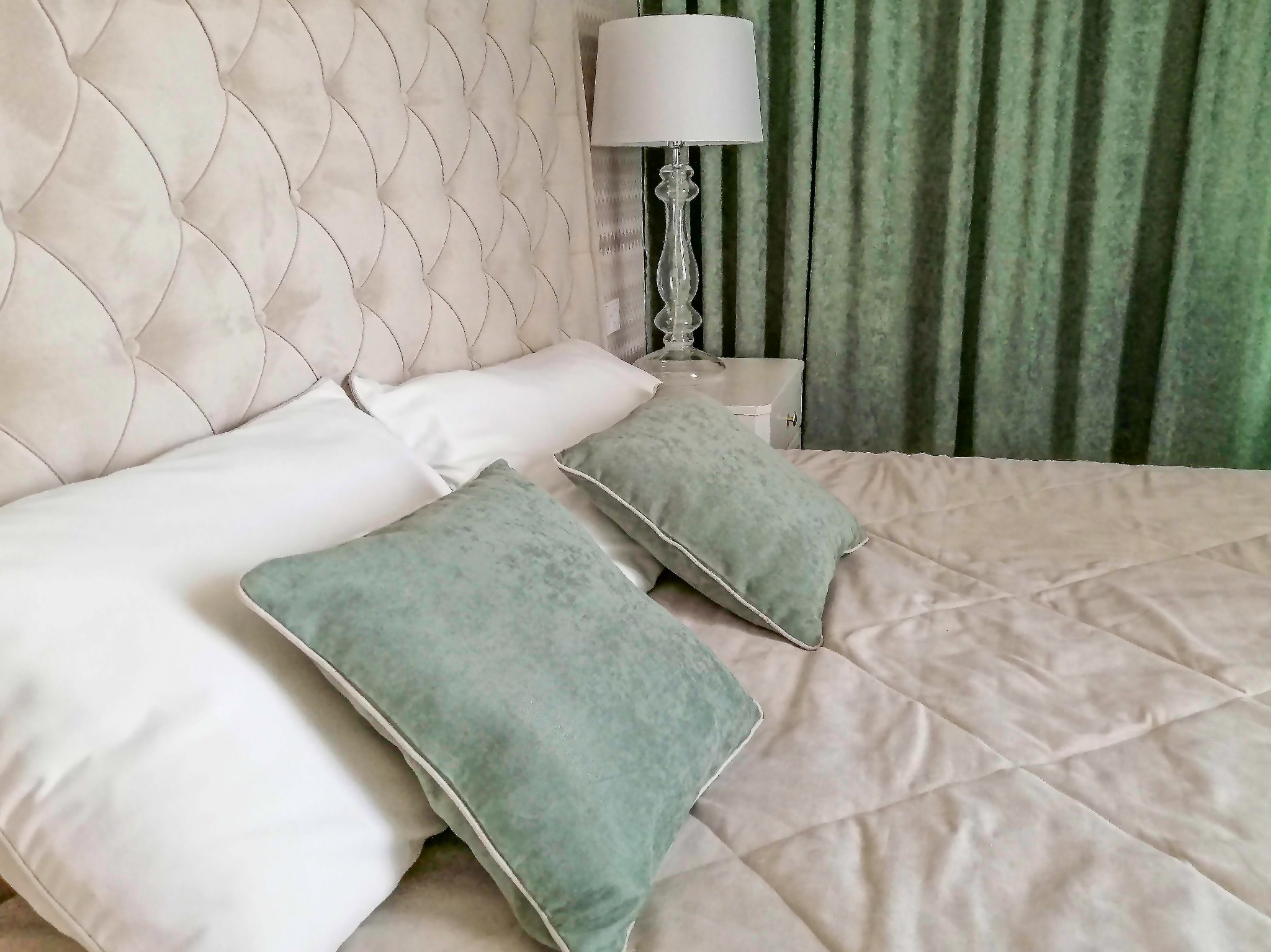 Beżowe pikowane wezgłowie łóżka z białymi poduszkami i dwoma zielonymi poduszkami dekoracyjnymi, obok lampka nocna z kryształową podstawą i zielone zasłony w tle.