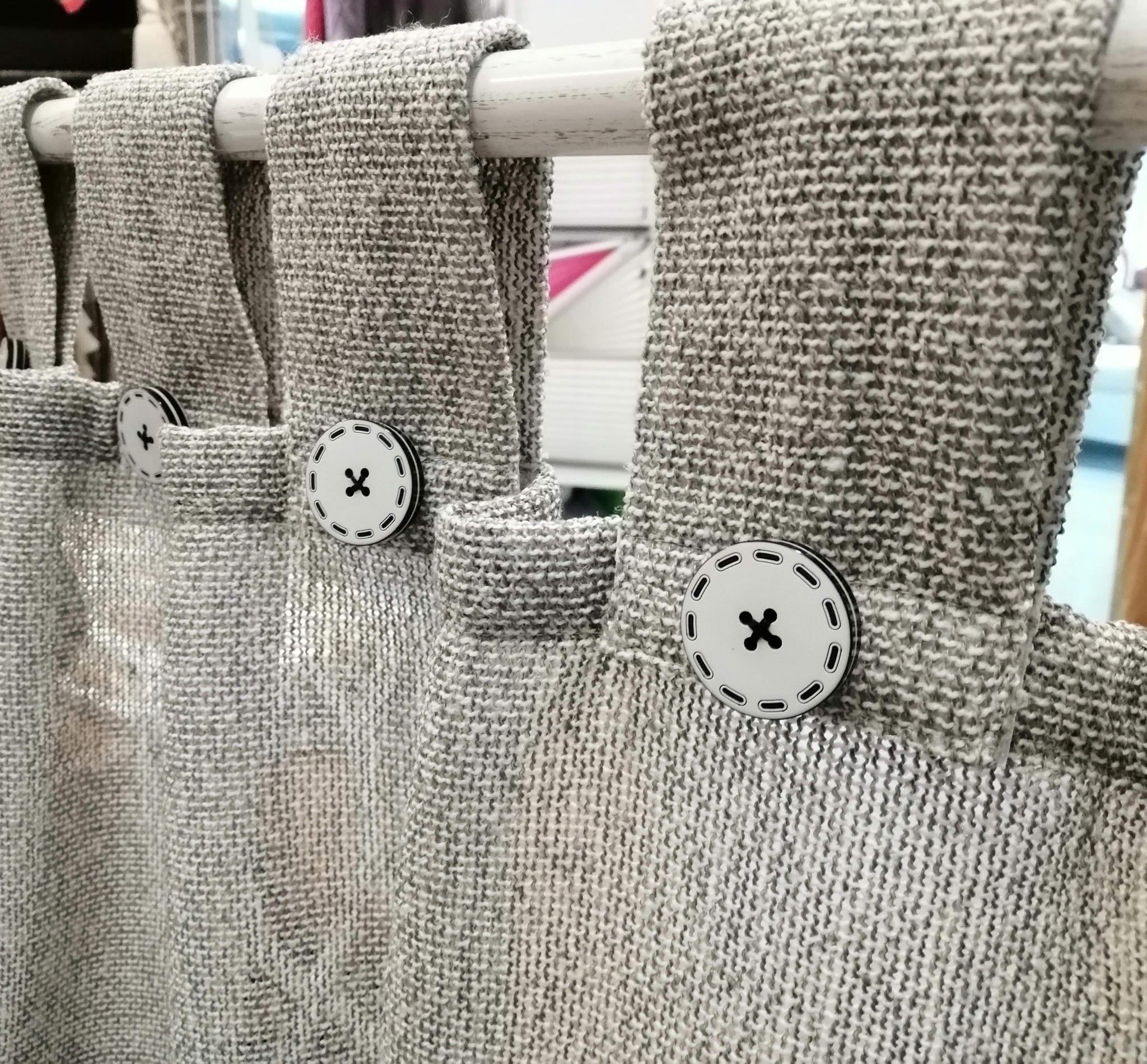 Fragment szarej, grubo tkanej zasłony z białymi guzikami z czarnym krzyżykiem, zawieszonej na białym karniszu.