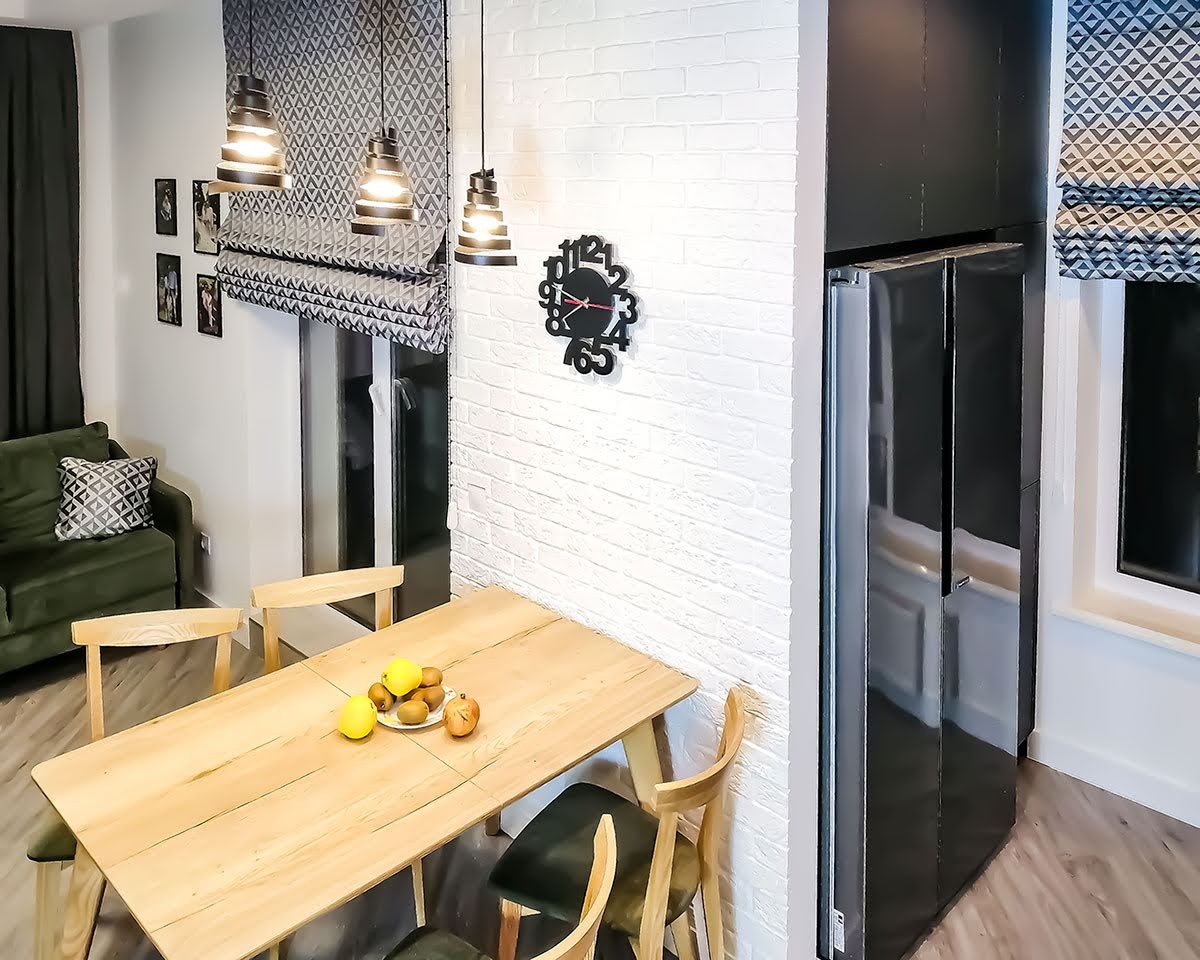 Jadalnia z drewnianym stołem, owocami na talerzu i roletami rzymskimi w geometryczny wzór, oświetlona trzema lampami wiszącymi.