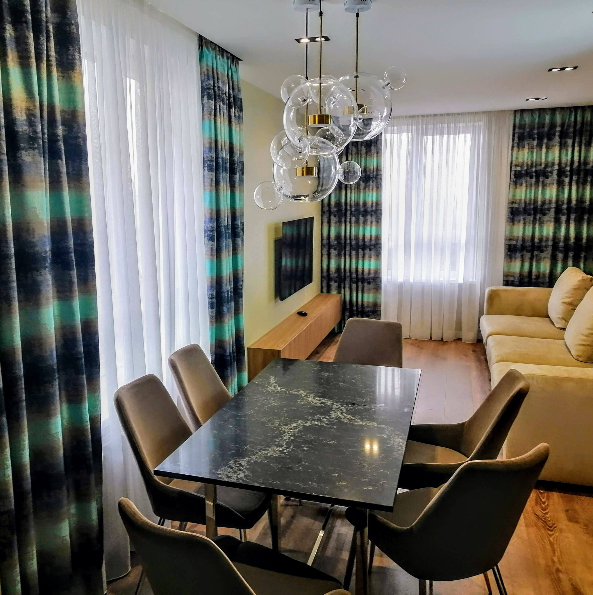 Salon z zasłonami w odcieniach turkusu, szarości i beżu, zestawionymi z białymi firanami. Wnętrze dopełnia czarny stół z marmurkowym wzorem, welurowe krzesła, nowoczesna lampa i beżowa sofa.