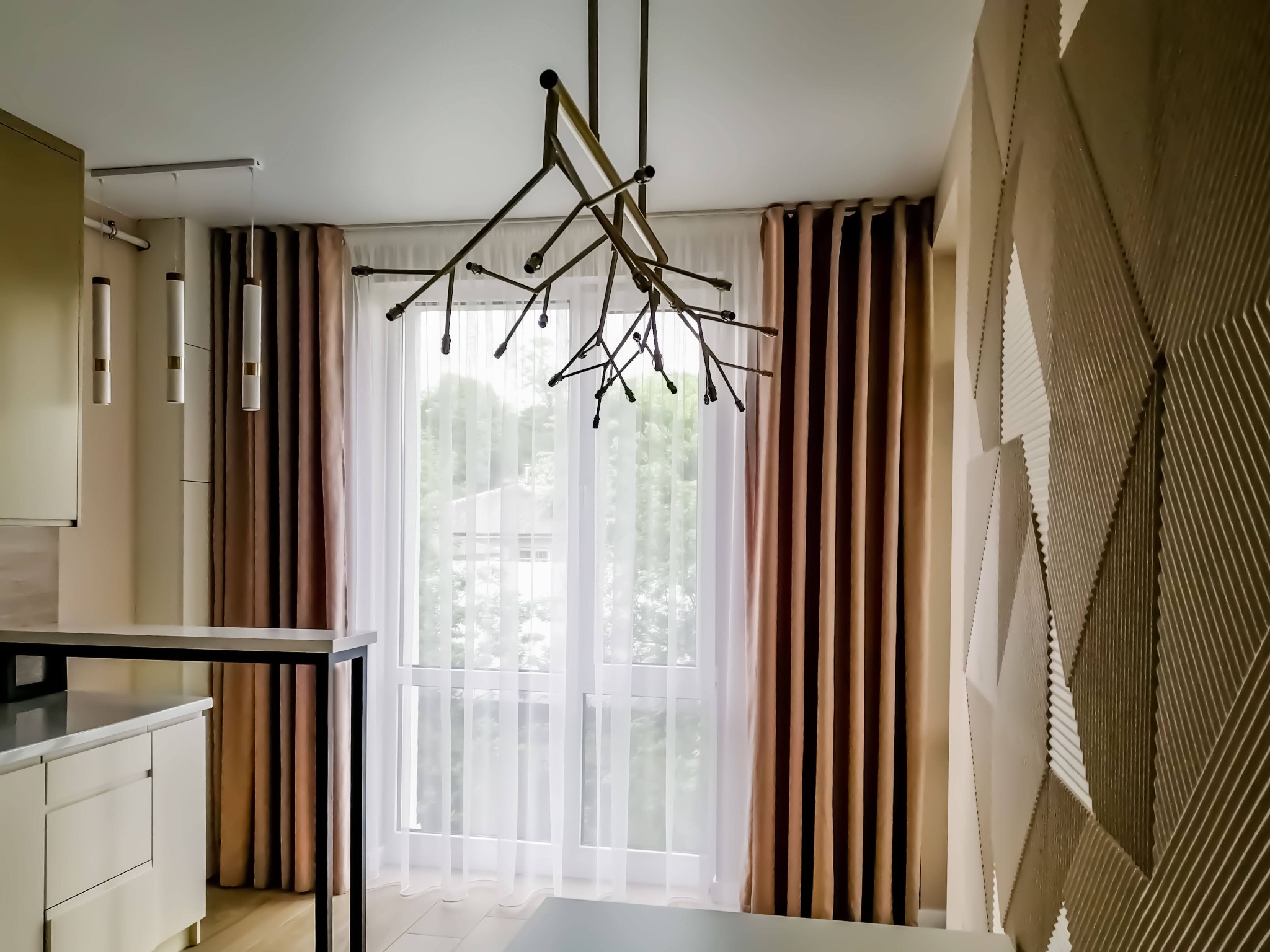 Nowoczesne wnętrze z beżowymi zasłonami, białymi firanami, designerską lampą sufitową i dekoracyjną ścianą z geometrycznym wzorem.