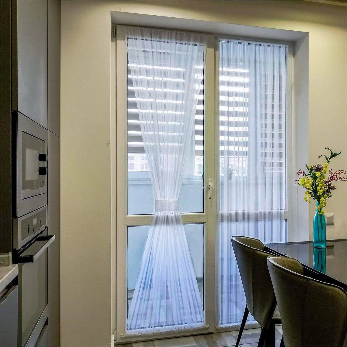 Delikatna, biała, transparentna firanka upięta w połowie wysokości ozdobną taśmą, zawieszona na białych drzwiach balkonowych w nowoczesnej kuchni z widokiem na zabudowania za oknem.