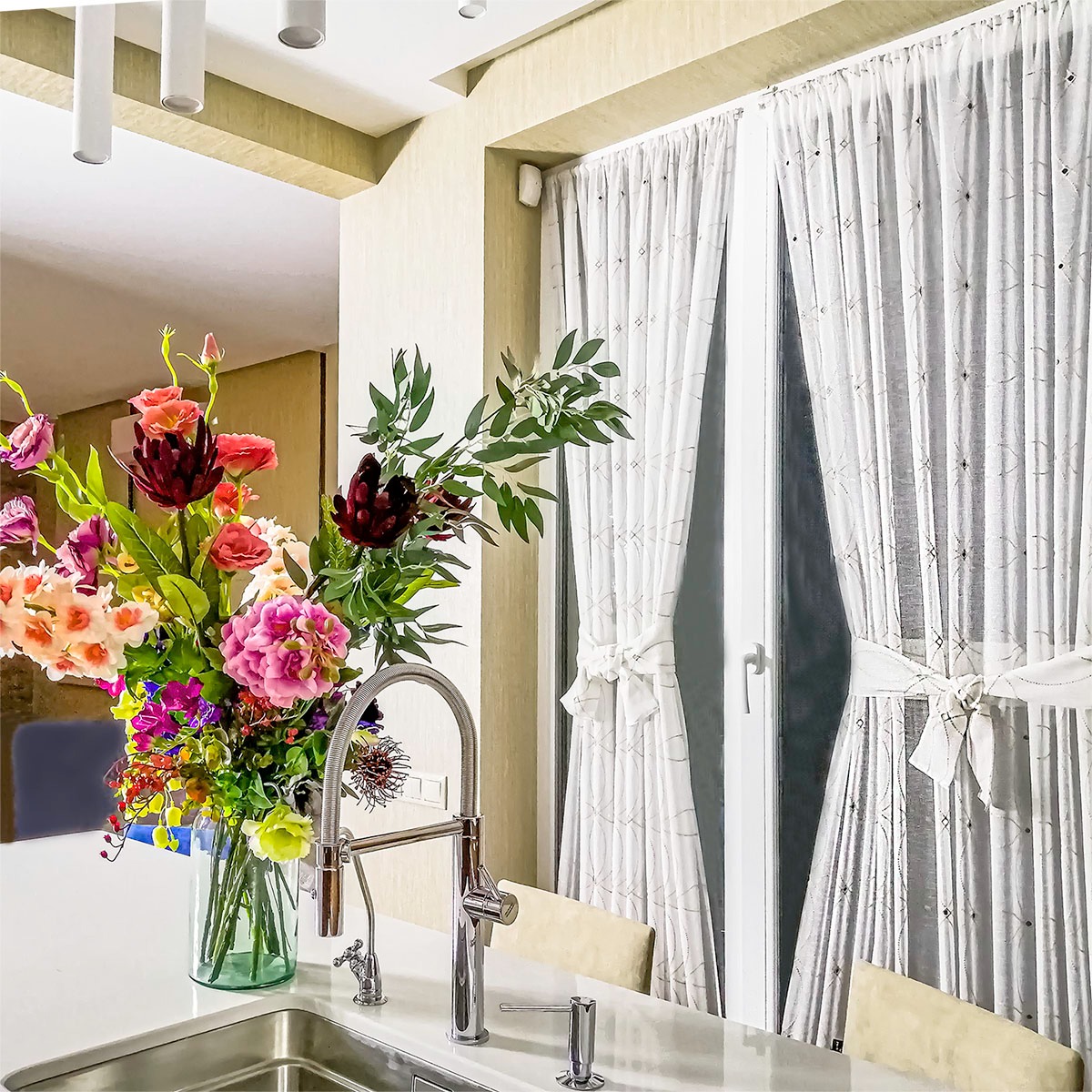 Jasne, białe zasłony z subtelnym wzorem, upięte kokardami, za nimi okno, na pierwszym planie bukiet kolorowych kwiatów w szklanym wazonie na blacie kuchennym z nowoczesną baterią.