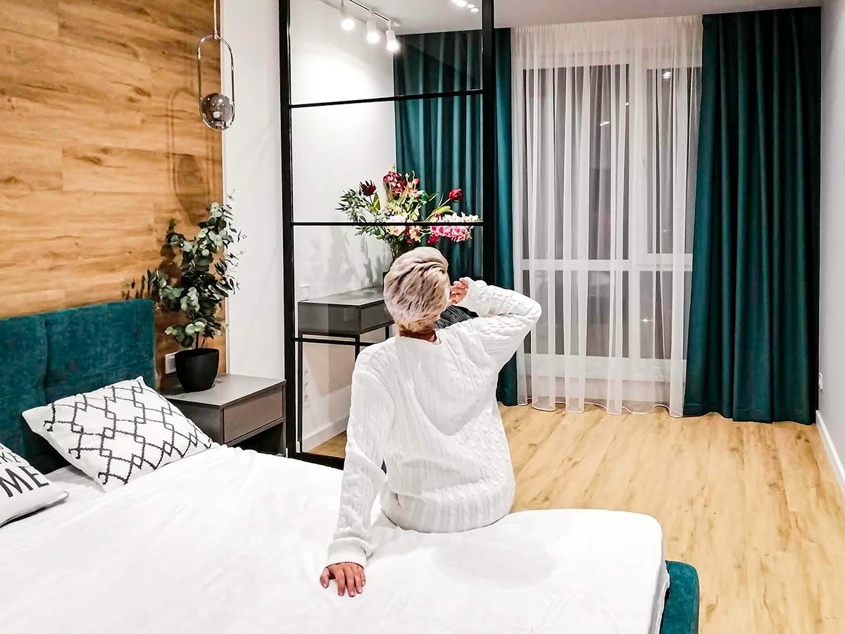 Jasna sypialnia z drewnianą ścianą, turkusowym zagłówkiem i zasłonami w odcieniach zieleni, kobieta w białym swetrze siedzi na łóżku tyłem do kamery.