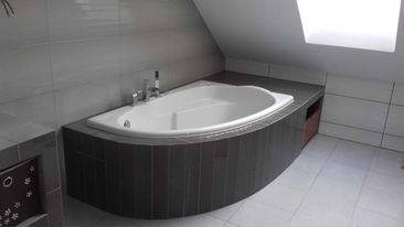 Biała wanna obudowana ciemnoszarą mozaiką w łazience na poddaszu ze skośnym sufitem i szarymi płytkami na ścianach.