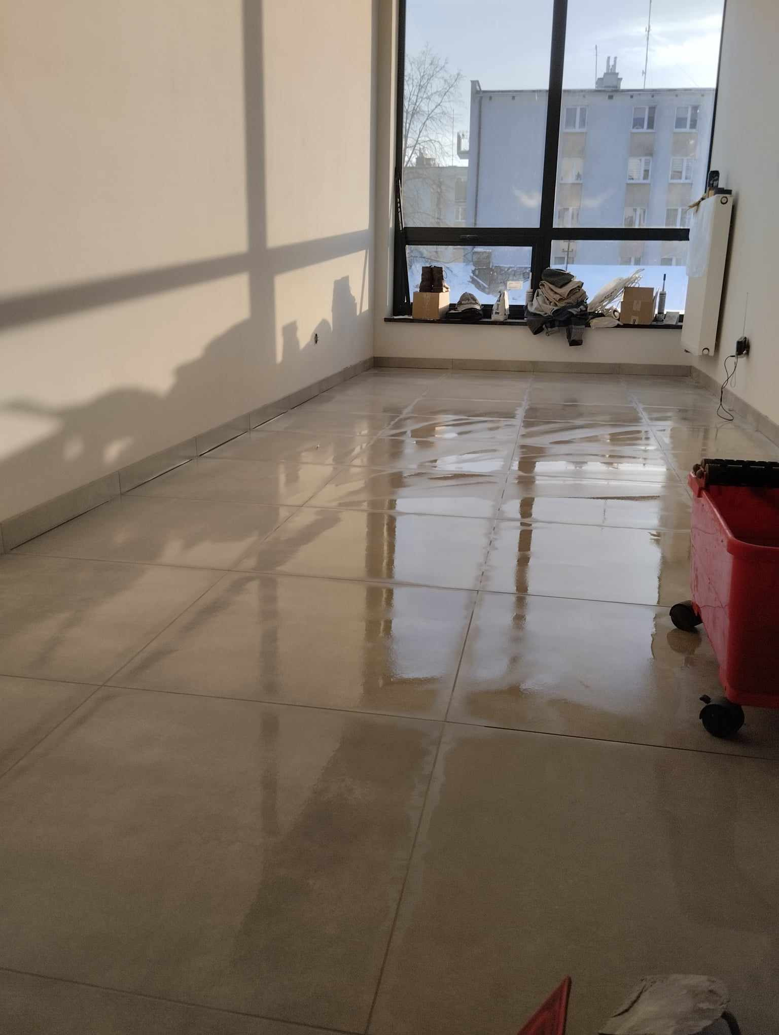 Świeżo położone, błyszczące płytki podłogowe w jasnym pomieszczeniu z dużym oknem. Widoczne narzędzia do fugowania i czerwone wiadro na kółkach.