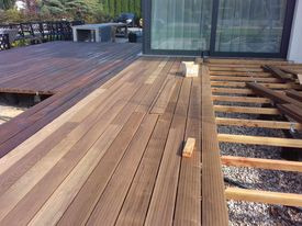 Montaż drewnianego tarasu z desek kompozytowych na zewnątrz domu. Część tarasu ukończona, widoczne deski w ciepłym odcieniu brązu. Druga część w trakcie budowy, ukazująca drewniany szkielet...