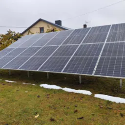 Instalacja paneli słonecznych na trawniku przed domem w pochmurny dzień. Widoczne resztki śniegu na ziemi. Ujęcie z bliska ukazuje strukturę paneli.