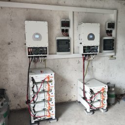 TSI Energy - Kompletna instalacja fotowoltaiczna w pomieszczeniu: dwa inwertery Deye, akumulatory i skrzynki z bezpiecznikami na tle surowej ściany. Widoczne przewody i butle gazowe.