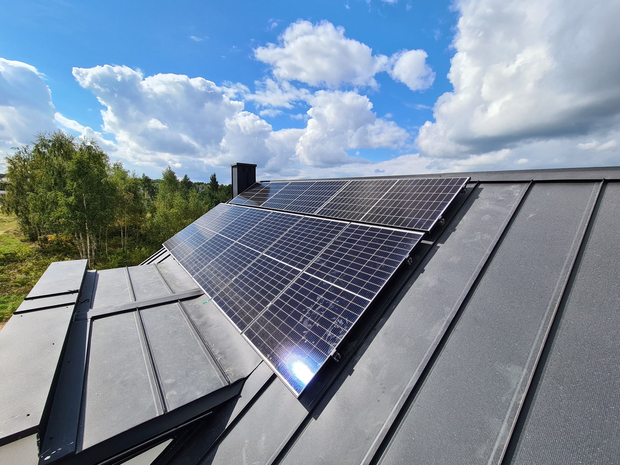 Panele słoneczne na szarym dachu blaszanym, widok z góry w słoneczny dzień. W tle drzewa i błękitne niebo z białymi chmurami.
