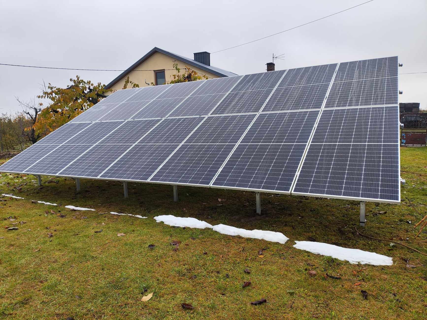 Instalacja paneli słonecznych na trawniku przed domem w pochmurny dzień. Widoczne resztki śniegu na ziemi. Ujęcie z bliska ukazuje strukturę paneli.
