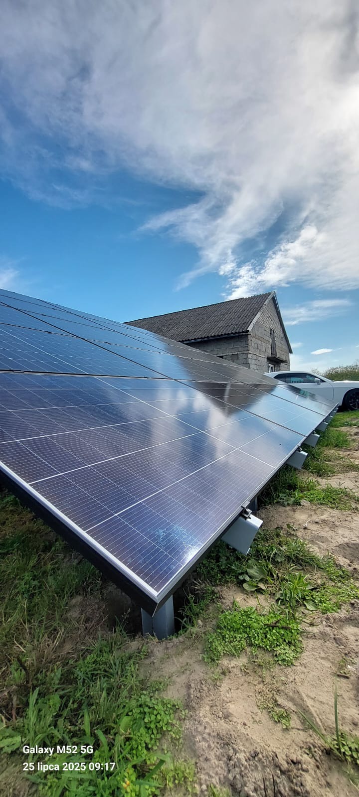 Instalacja fotowoltaiczna na gruncie z widokiem na budynek gospodarczy i samochód w tle, błękitne niebo z chmurami odbija się w panelach.