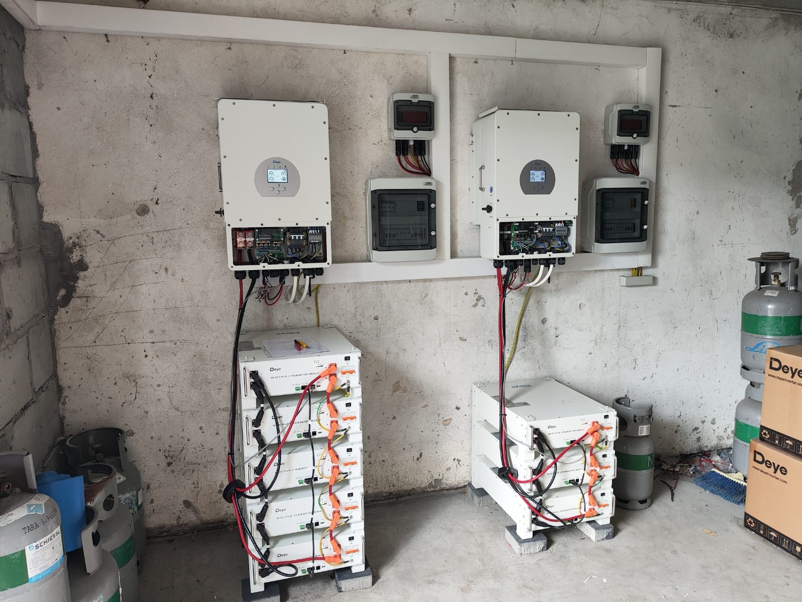 Kompletna instalacja fotowoltaiczna w pomieszczeniu: dwa inwertery Deye, akumulatory i skrzynki z bezpiecznikami na tle surowej ściany. Widoczne przewody i butle gazowe.