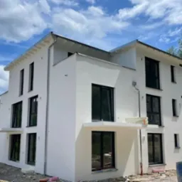 Nowoczesny, dwupiętrowy dom z białymi elewacjami i czarnymi ramami okiennymi, widoczny z zewnątrz na tle błękitnego nieba z chmurami. Wokół budynku widoczne materiały budowlane.