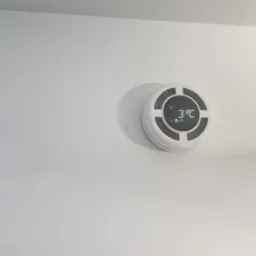 Biały, okrągły rekuperator ścienny z wyświetlaczem pokazującym temperaturę 3°C, zamontowany na gładkiej, białej ścianie.