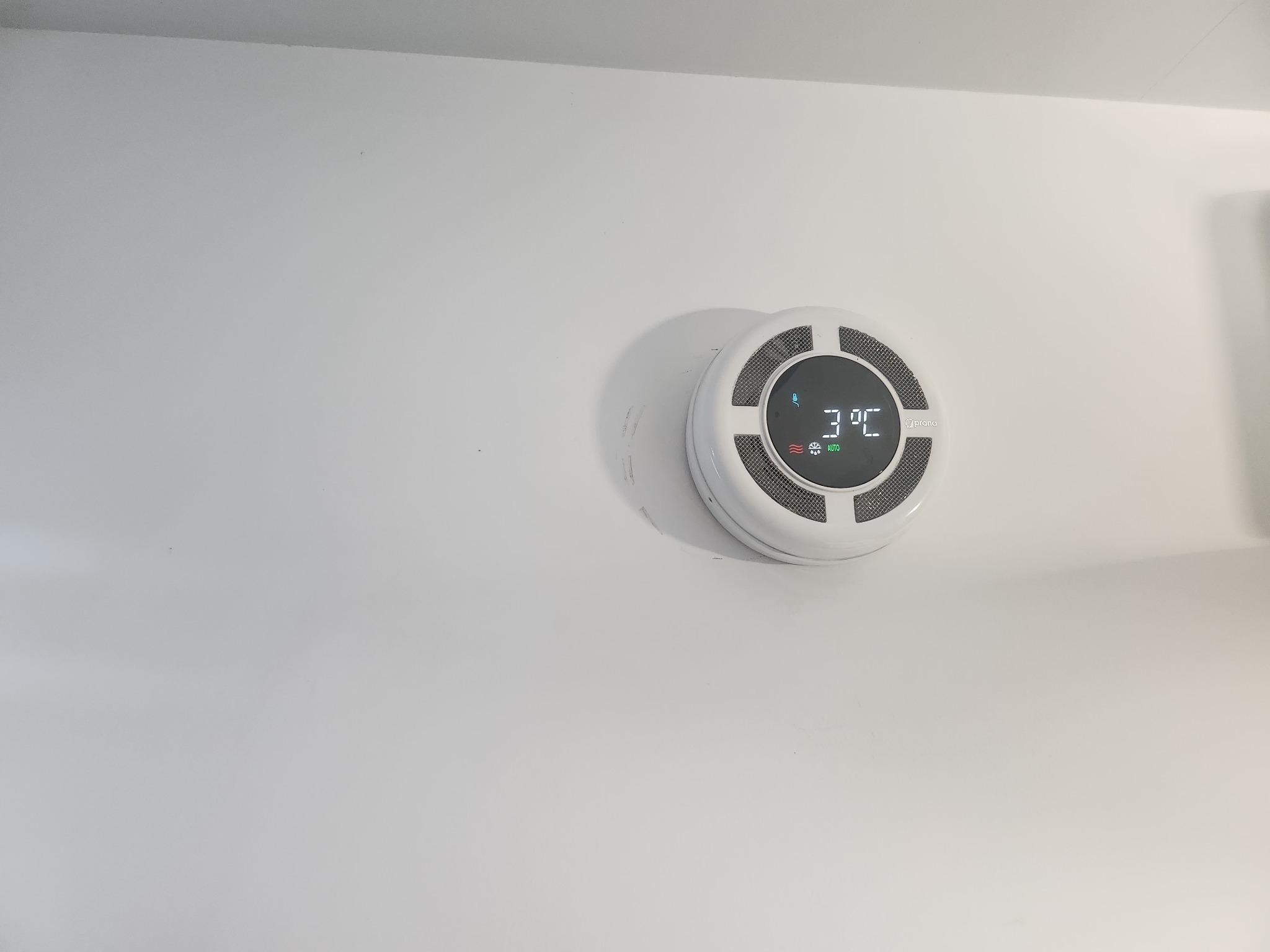 Biały, okrągły rekuperator ścienny z wyświetlaczem pokazującym temperaturę 3°C, zamontowany na gładkiej, białej ścianie.