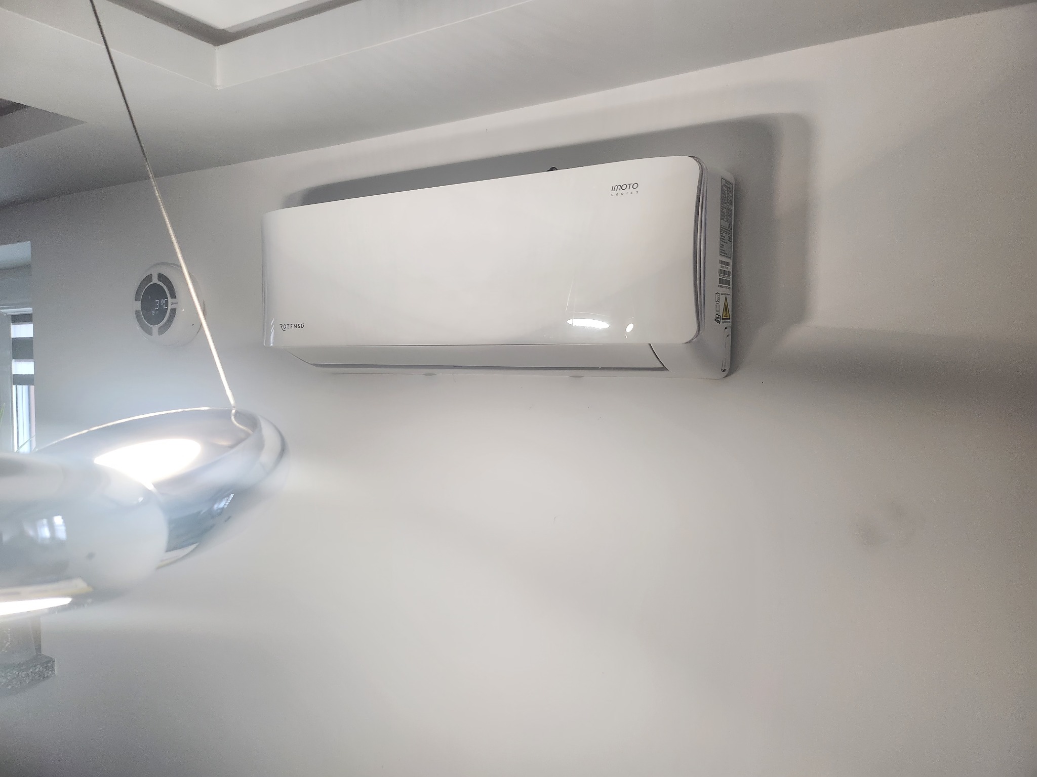 Biały klimatyzator marki Imoto Series zamontowany na ścianie w pomieszczeniu z widocznym fragmentem lampy i okrągłym czujnikiem temperatury.