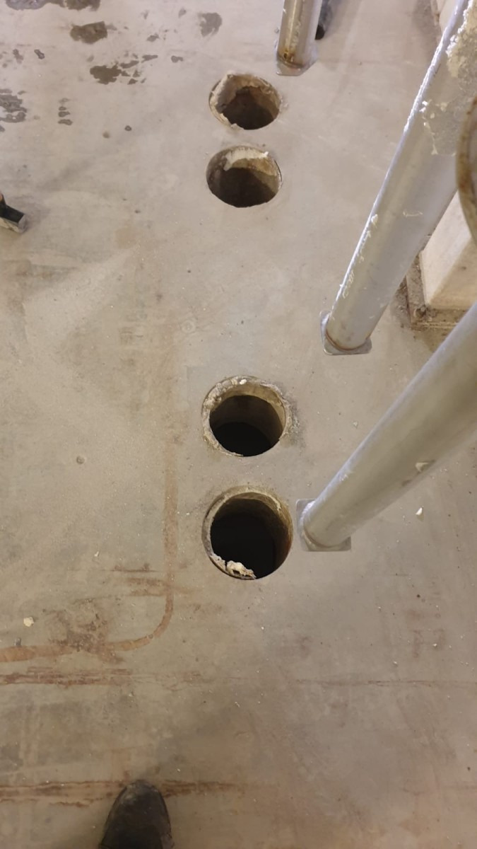 Betonowa podłoga z wywierconymi otworami, widoczne fragmenty stalowej konstrukcji i but pracownika w rogu kadru.