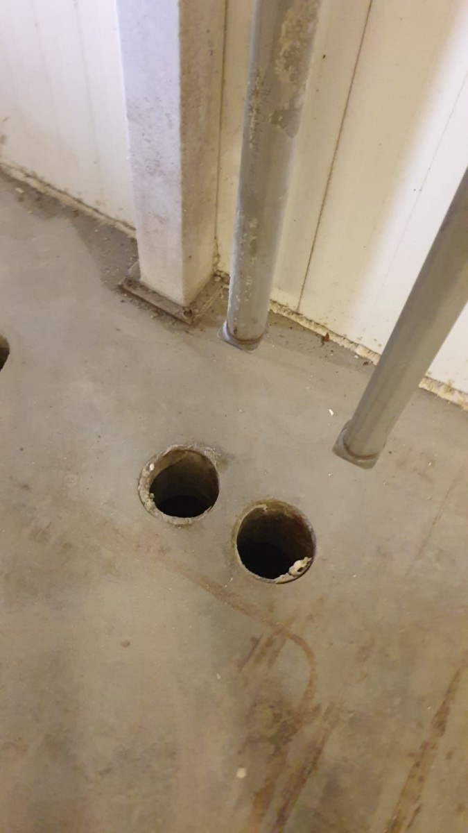 Dwa okrągłe otwory wywiercone w betonowej podłodze, widoczne fragmenty rur instalacyjnych przy ścianie.