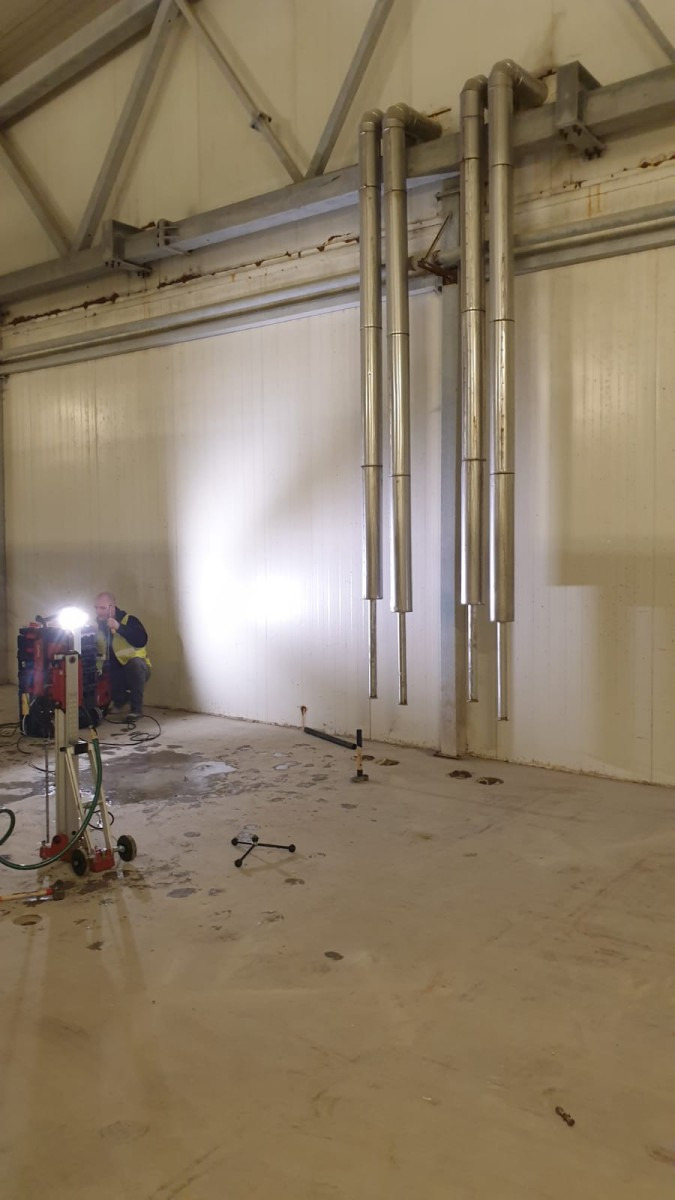 Montaż rur wentylacyjnych w hali przemysłowej, widoczny pracownik z wiertnicą diamentową i wywiercone otwory w posadzce.