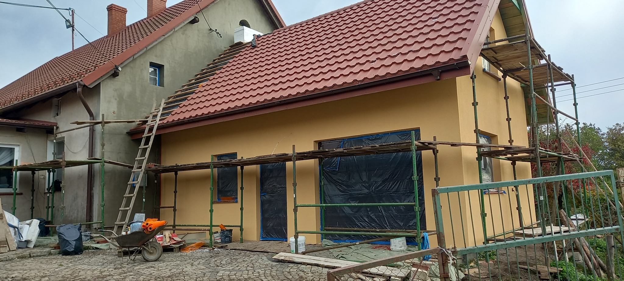 Dom jednorodzinny w trakcie malowania elewacji na żółto, z widocznymi rusztowaniami, zabezpieczonymi oknami i częściowo wymienionym dachem z czerwonej dachówki. W tle drugi budynek z szarą elewacją.