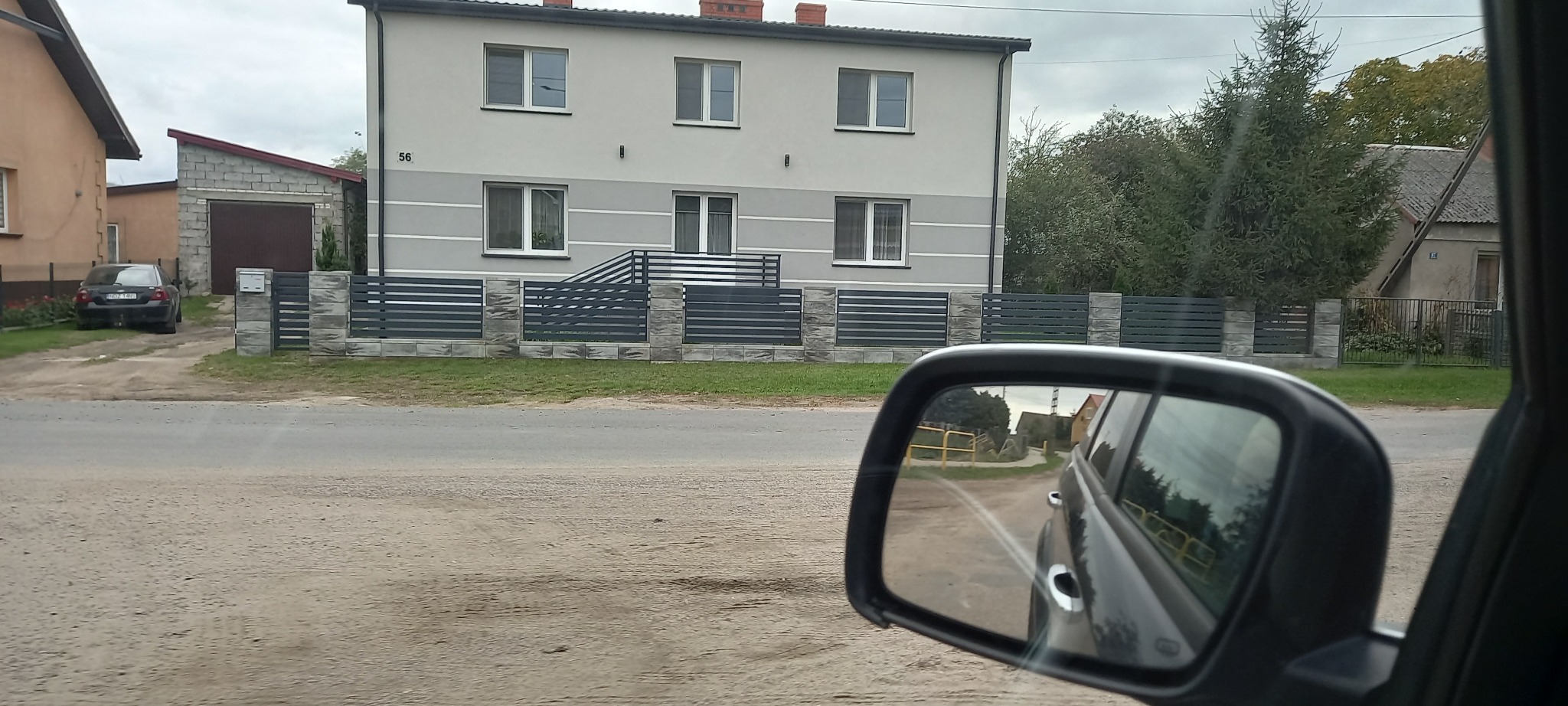 Dwukondygnacyjny dom z szarą elewacją i ciemnoszarym, poziomym ogrodzeniem, widziany z wnętrza samochodu poprzez lusterko boczne.