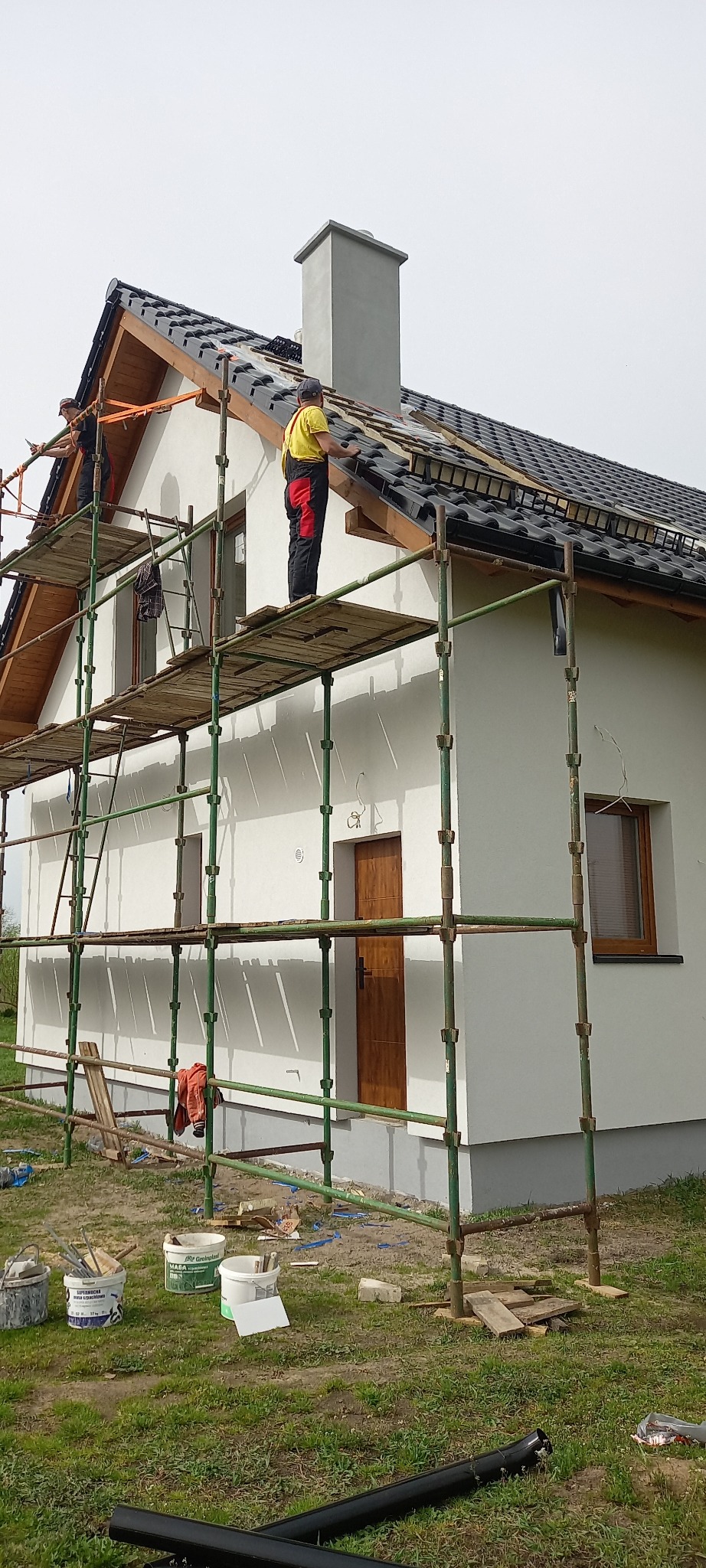 Prace dekarskie na dachu domu jednorodzinnego z białymi ścianami, widok z rusztowania na montaż czarnej dachówki, w tle komin i pochmurne niebo.