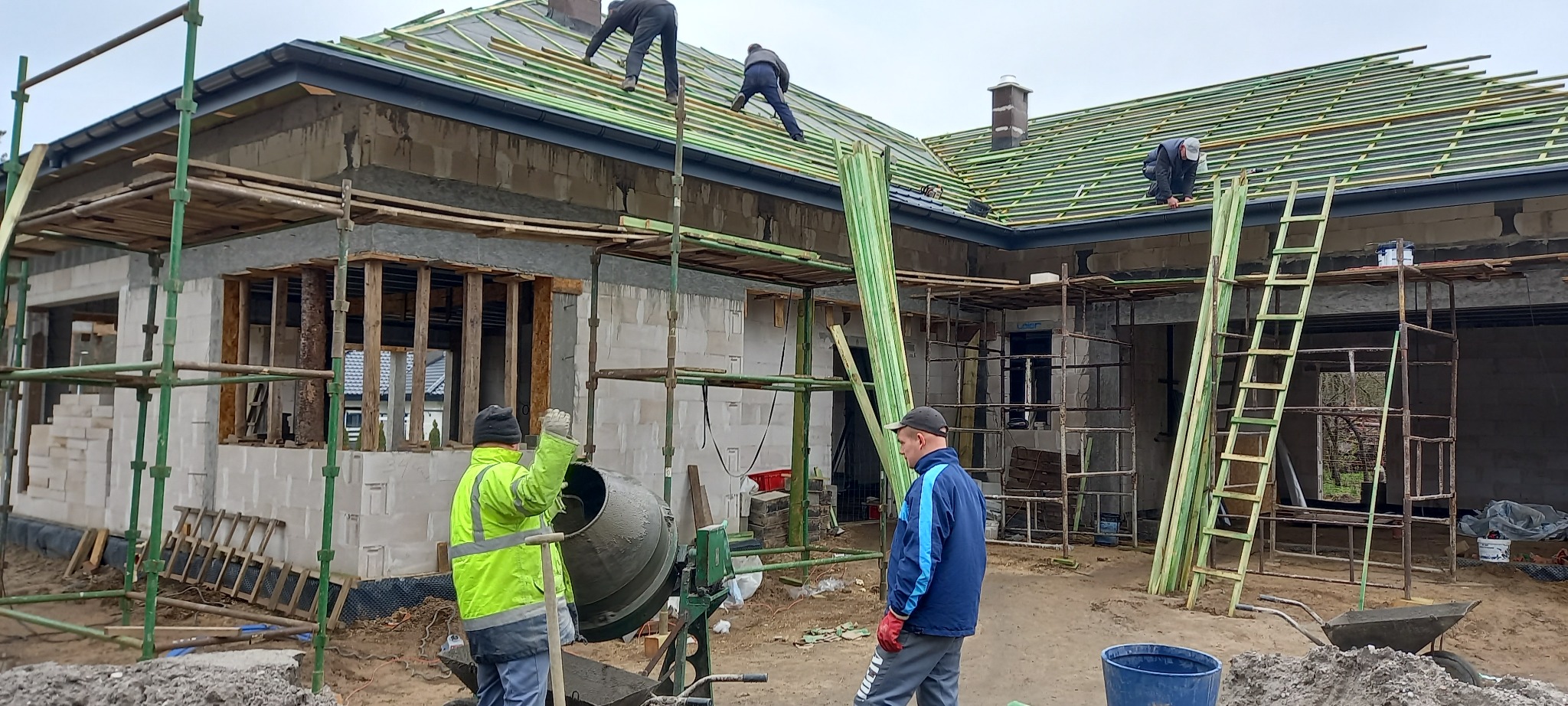 Budowa domu w trakcie realizacji: murowane ściany z betonu komórkowego, montaż drewnianej konstrukcji dachu, betoniarka i pracownicy na budowie.