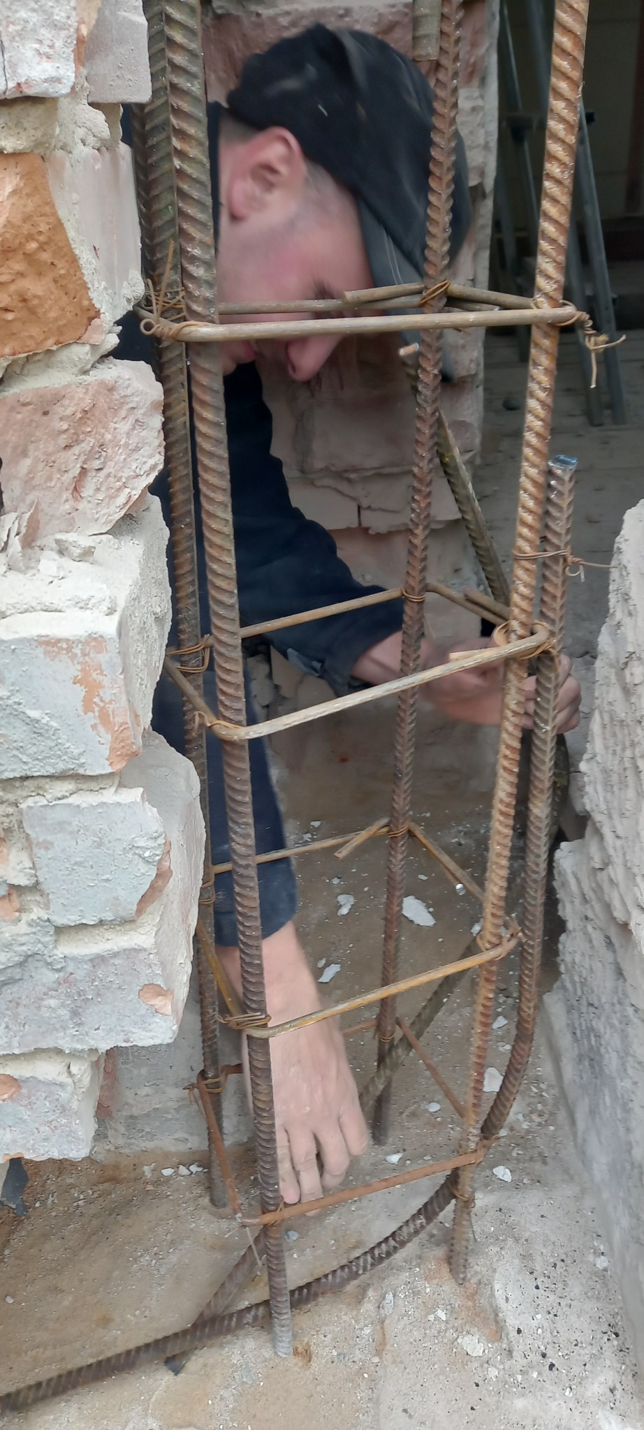 Zbrojenie kolumny z betonu zbrojonego w trakcie budowy, widoczne pręty zbrojeniowe połączone drutem, fragment ściany z cegły i betonowa wylewka.