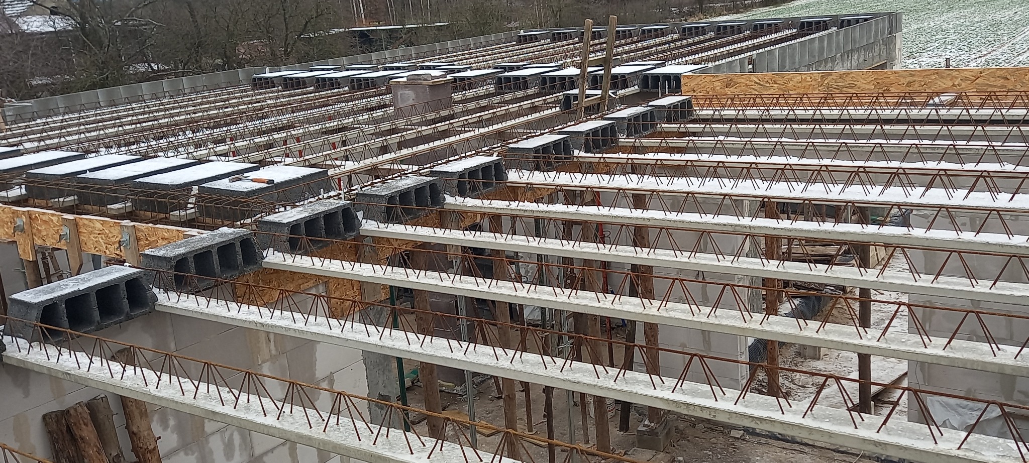 Wczesny etap budowy domu z widocznymi betonowymi belkami stropowymi, bloczkami betonowymi i zbrojeniami, lekko przyprószone śniegiem.