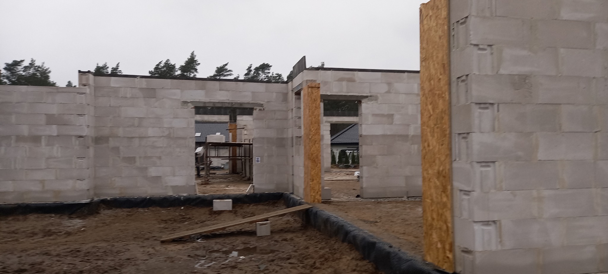 Surowy stan budynku z betonu komórkowego w trakcie wznoszenia, widoczne otwory okienne i drzwiowe, grunt wewnątrz zabezpieczony folią, drzewa w tle.