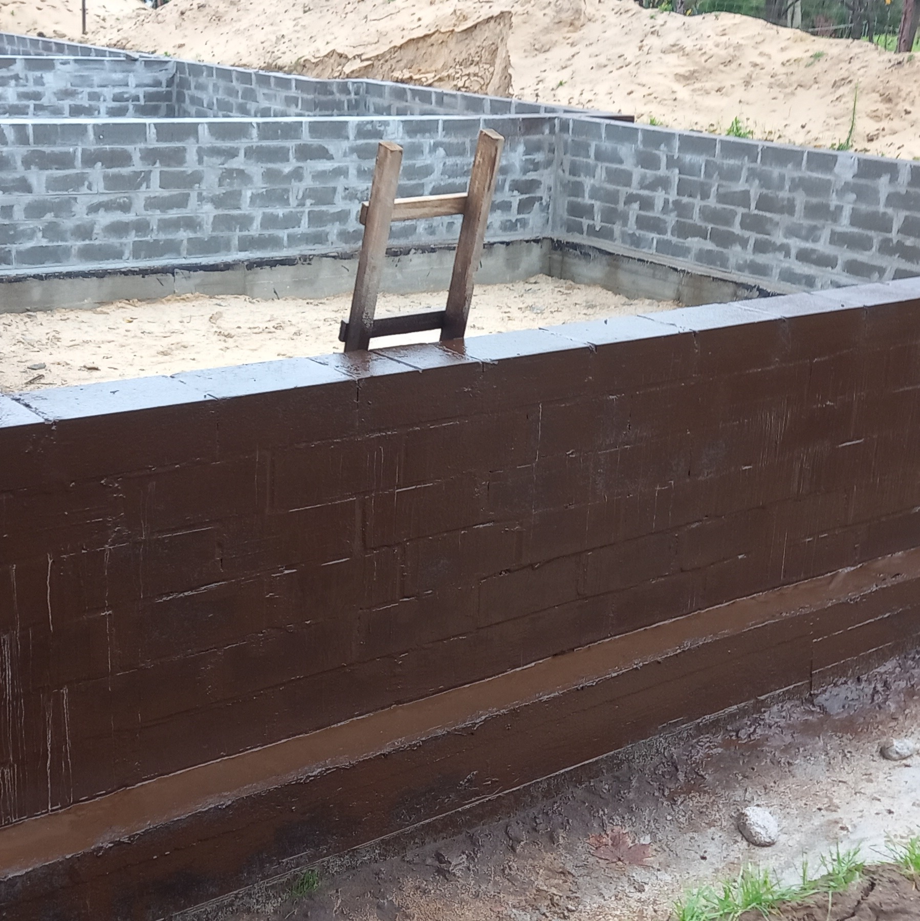Zabezpieczone, brązową izolacją, ściany fundamentowe z bloczków betonowych, widoczna drabina drewniana i piasek wewnątrz wykopu.