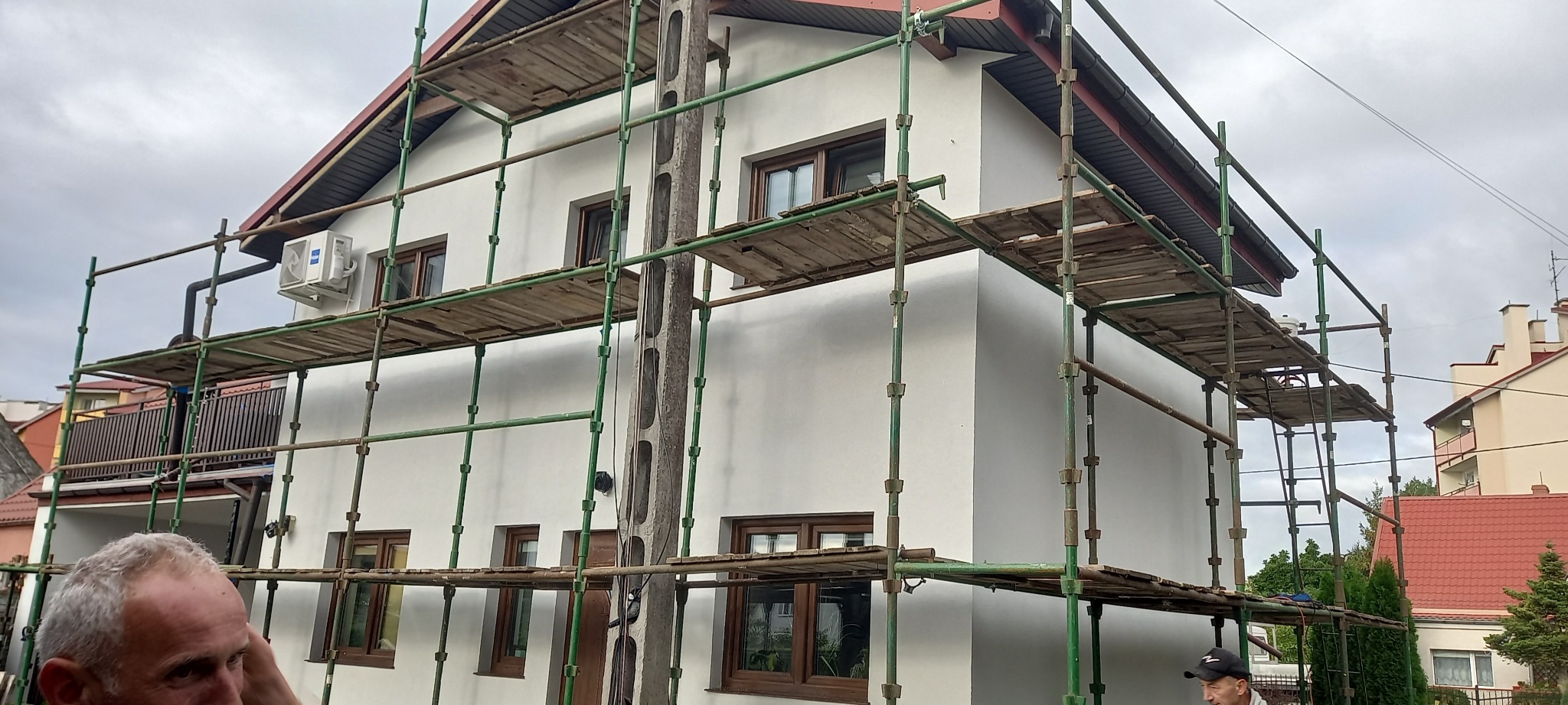 Biały dom jednorodzinny w trakcie prac elewacyjnych, otoczony rusztowaniem, z widocznym klimatyzatorem na ścianie i częściowo zasłoniętą twarzą mężczyzny w lewym dolnym rogu.