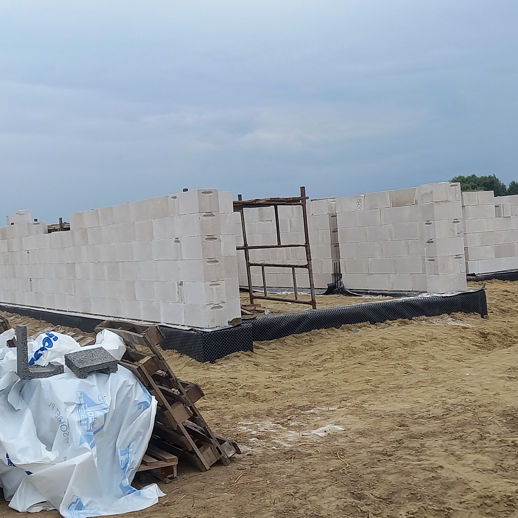 Wznoszenie ścian parteru z bloczków betonu komórkowego na przygotowanym fundamencie z hydroizolacją, widoczne rusztowanie i materiały budowlane na placu budowy.