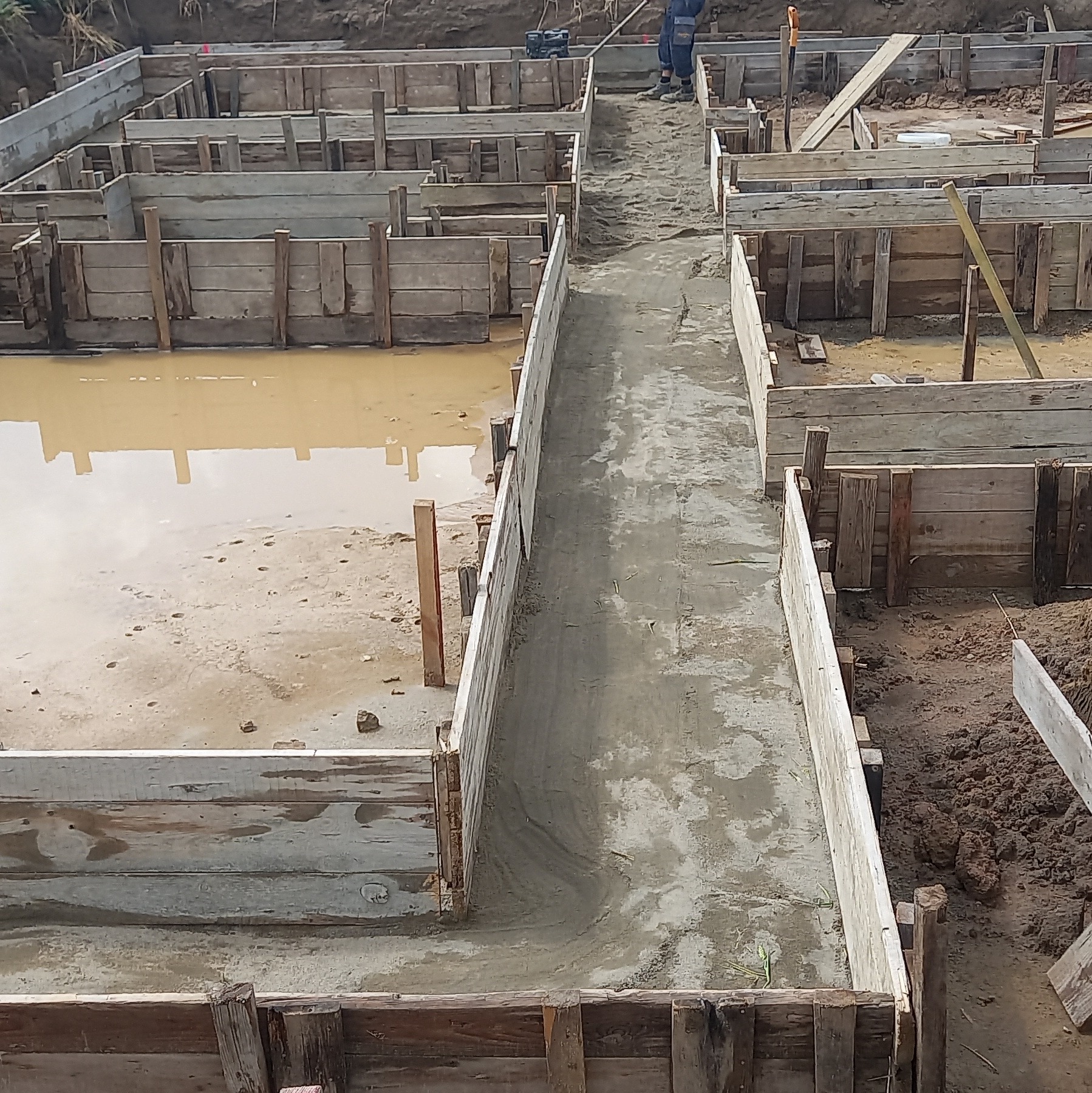 Zalewanie betonem szalunków fundamentowych z drewnianych desek, widoczne ślady wody na ziemi i narzędzia w tle.
