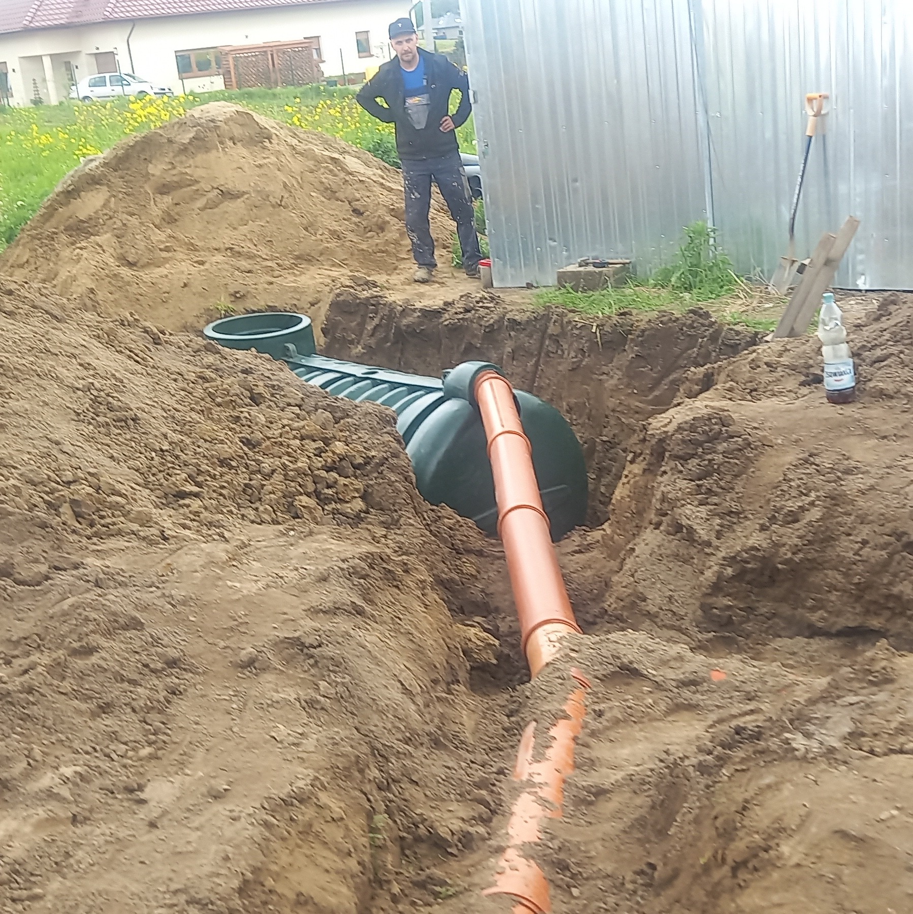 Montaż szamba ekologicznego z rurami odprowadzającymi w wykopie ziemnym, widoczny pracownik w tle, obok sterta ziemi i butelka wody.