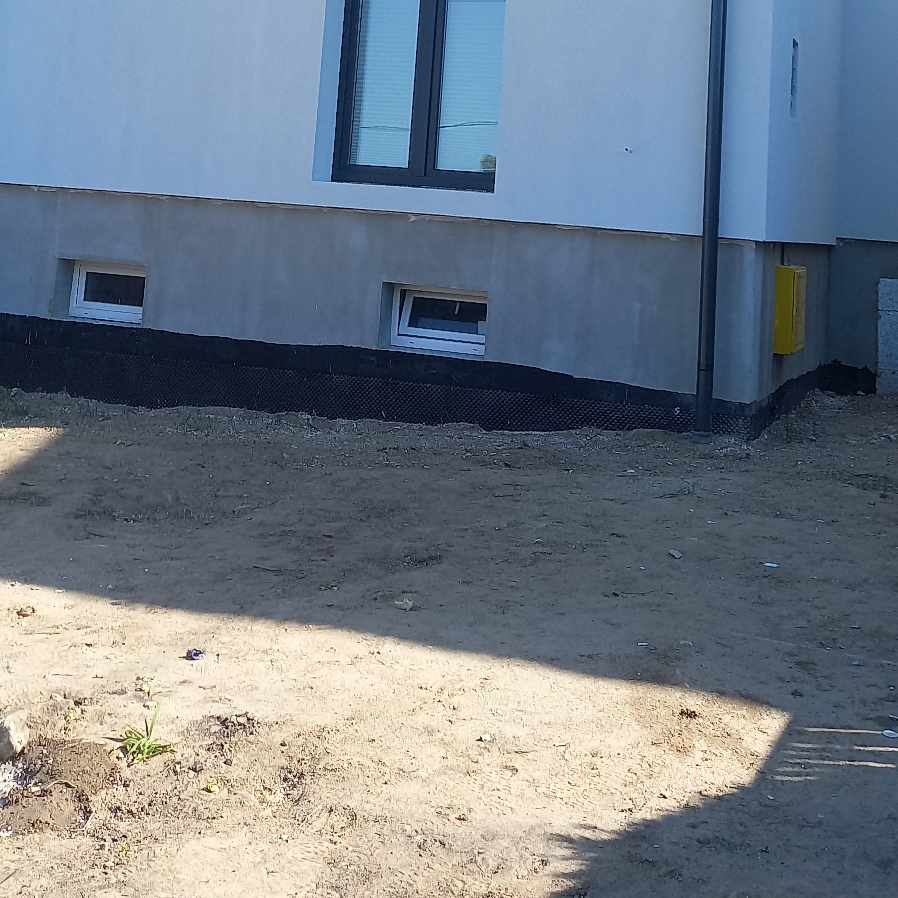 Fragment nowo wybudowanego domu z widocznymi oknami, fundamentem zabezpieczonym folią kubełkową i przyłączem gazowym na ścianie zewnętrznej.