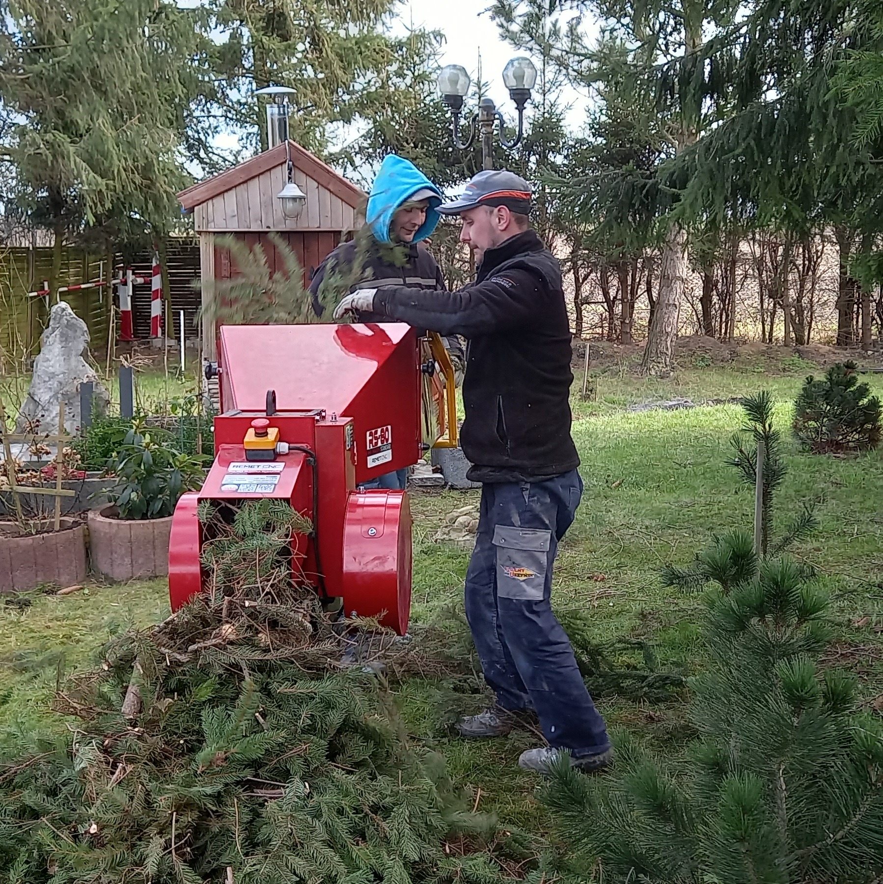 Dwóch mężczyzn obsługuje czerwoną maszynę do rozdrabniania gałęzi w ogrodzie, w tle widoczny mały drewniany domek narzędziowy i drzewa.