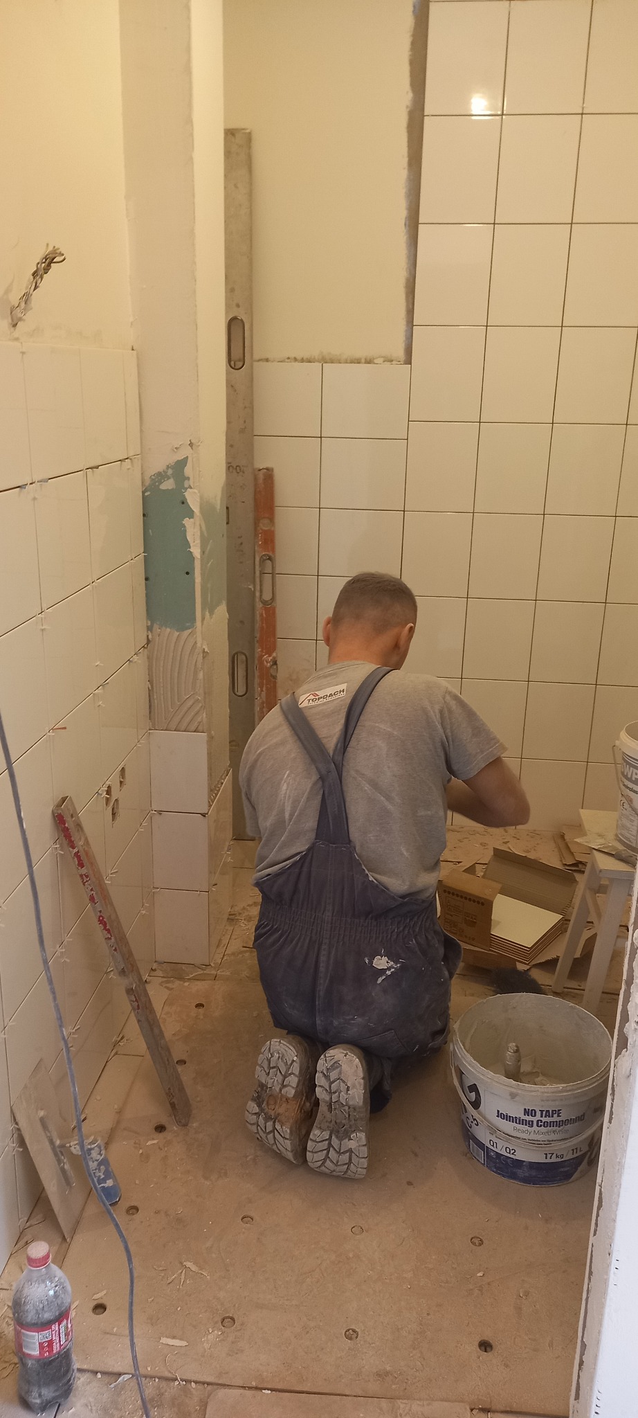 Pracownik w kombinezonie układa białe płytki w łazience, widoczne poziomice, wiadro z zaprawą i niedokończone fragmenty ścian.