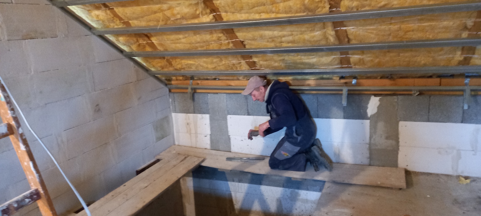 Pracownik na kolanach przy ścianie z bloków, montuje białe płyty izolacyjne, widoczne drewniane deski na podłodze i ocieplenie sufitu z metalowymi profilami