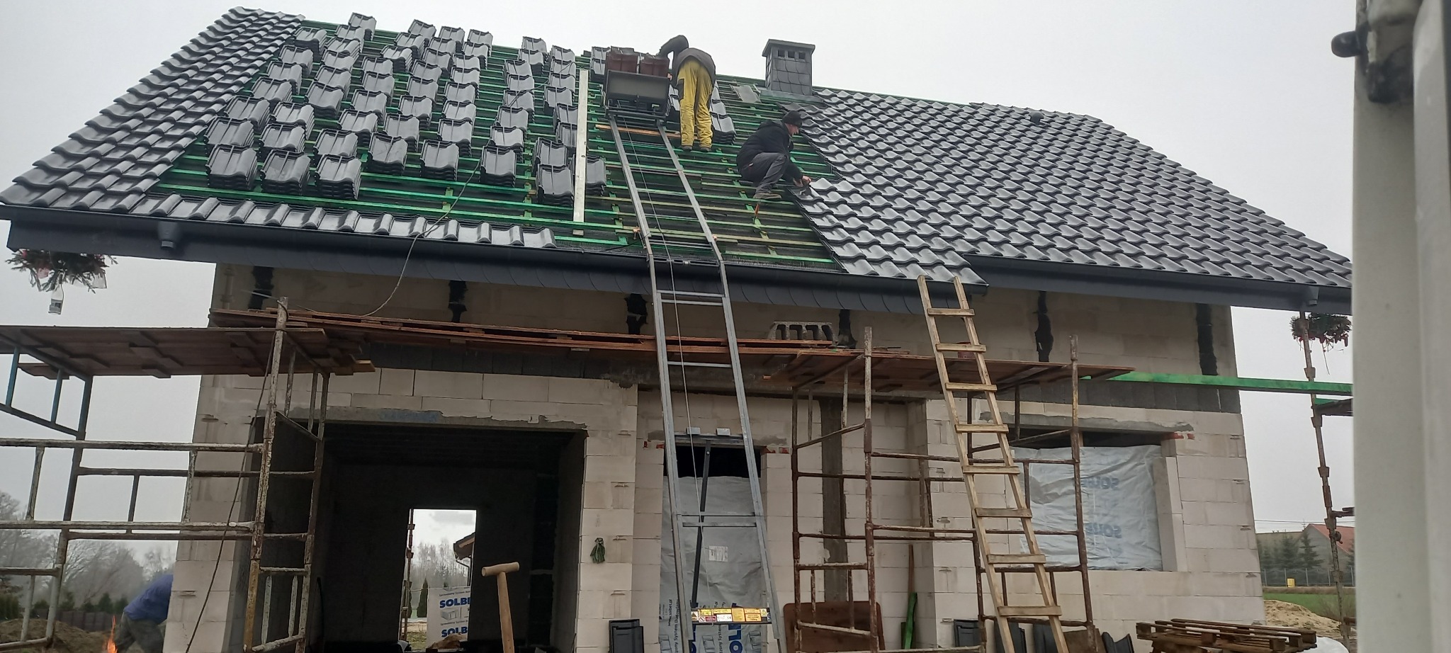 Budowa domu jednorodzinnego: dekarze układają czarną dachówkę na zielonej membranie, widoczne rusztowania i drabiny, stan surowy otwarty.